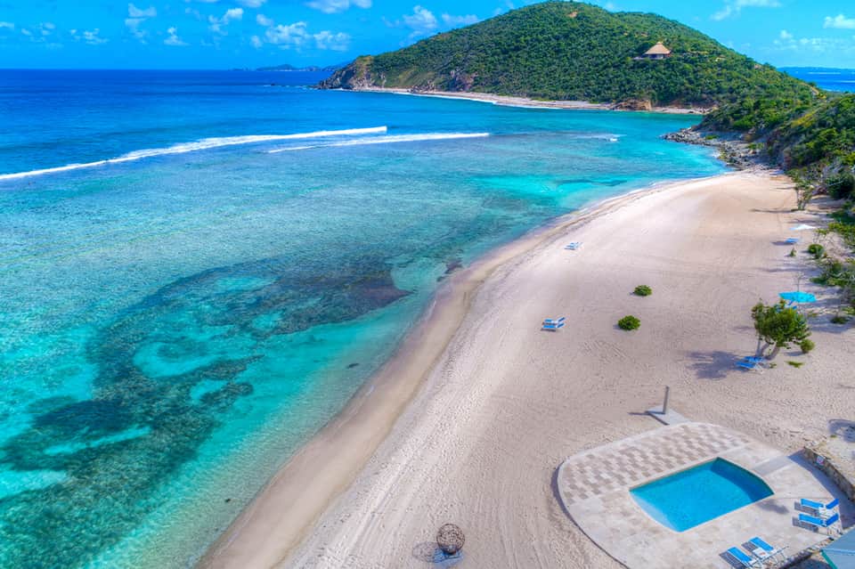 Pristine tropical beach with turquoise waters, white sand, forested hillside, and resort pool area