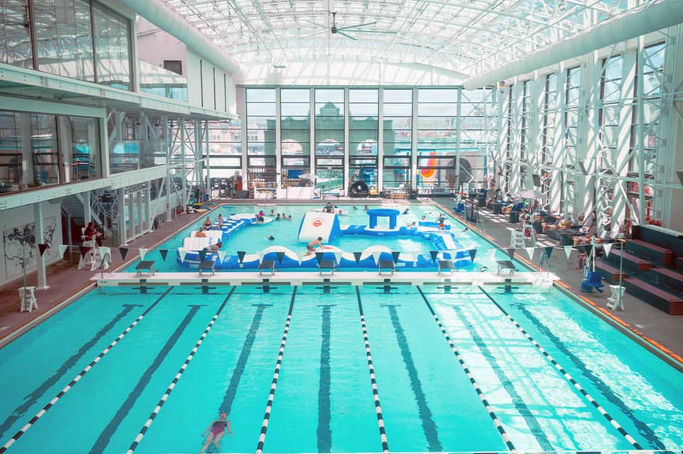 Olympic-size indoor swimming pool with lap lanes, diving board, spectator seating, and expansive glass roof structure