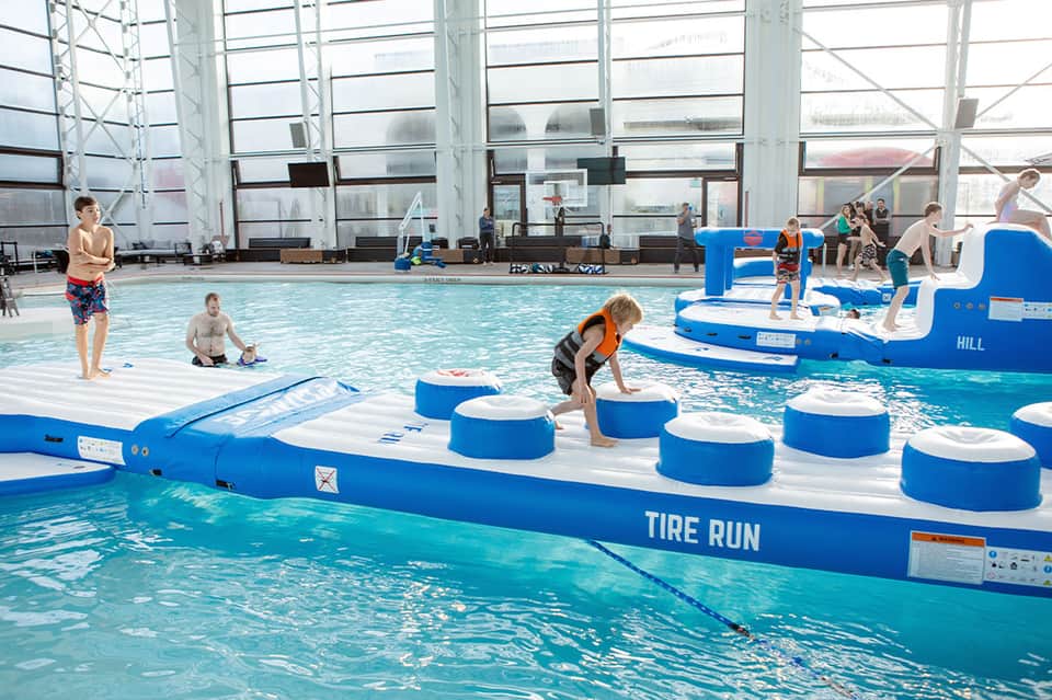 Indoor aquatic center with activity obstacles, children playing in shallow water, and glass ceiling