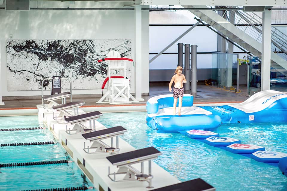 Indoor aquatic center with lap pool, diving board, inflatable play features, and young child on blue float