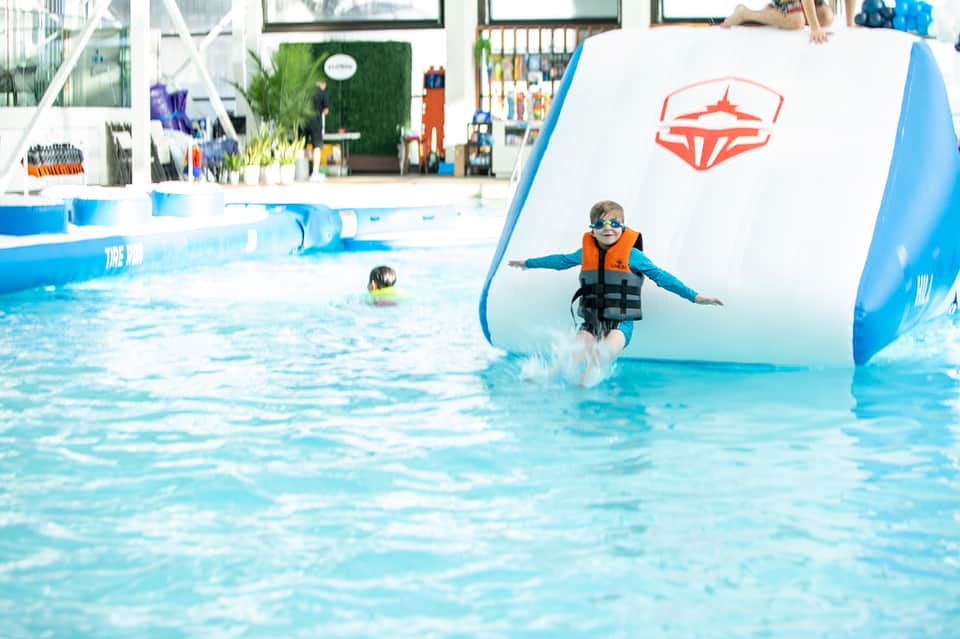 Child in life jacket splashing into indoor pool with inflatable obstacle course