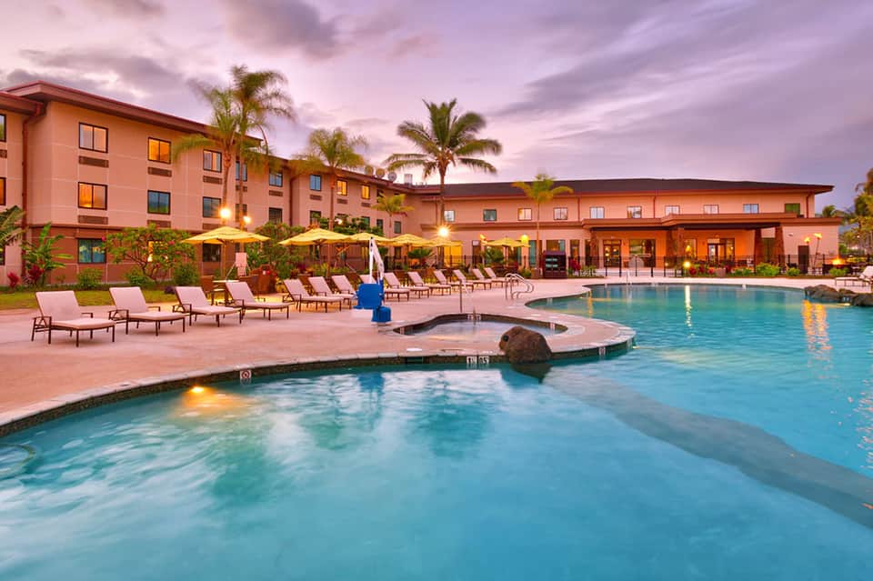 Resort pool at dusk with orange building, lounge chairs, and palm trees lit at night