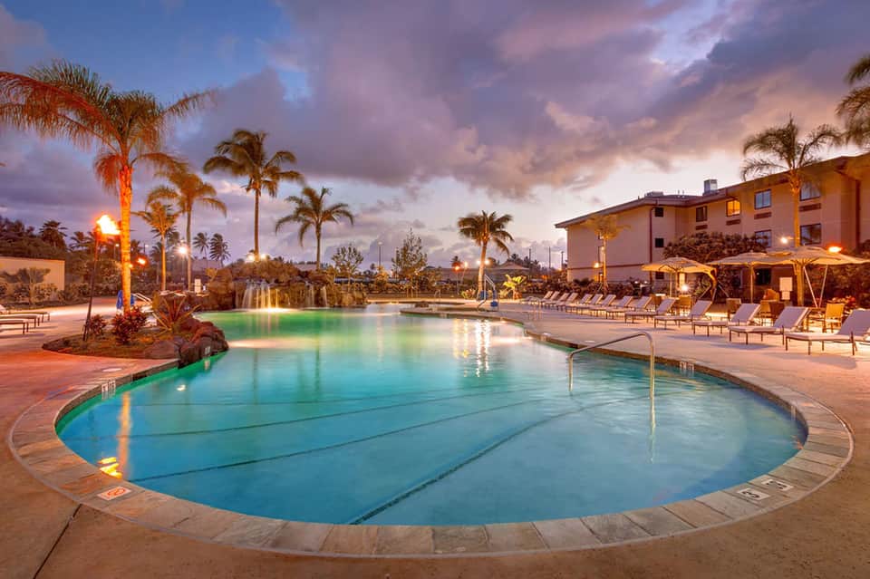 Illuminated resort pool at dusk with palm trees, lounge chairs, and warm lighting