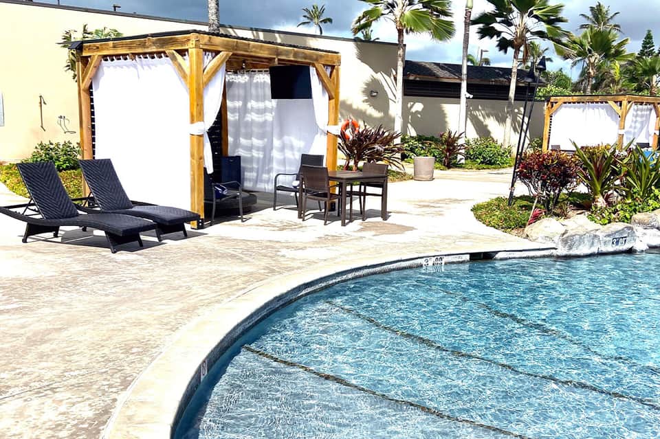 Pool area with wooden pergola cabana, loungers, tropical landscaping, and calm water