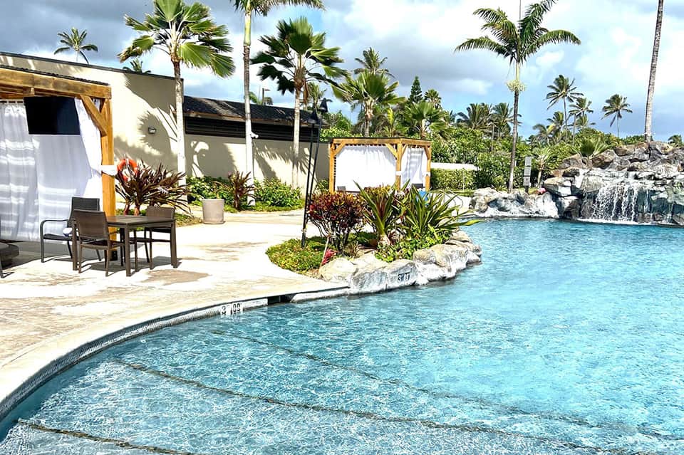 Lagoon-style resort pool with white cabanas, wooden pergolas, palms, and rock features