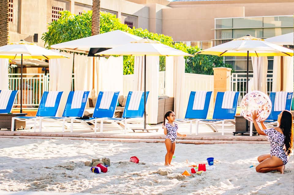 Children playing beach ball on sand with blue and white striped lounge chairs and yellow umbrellas in background
