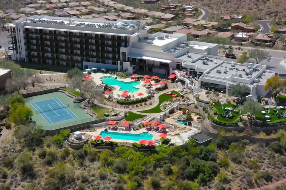 Aerial view of modern resort with turquoise pools, red umbrellas, tennis court, and surrounding desert landscape