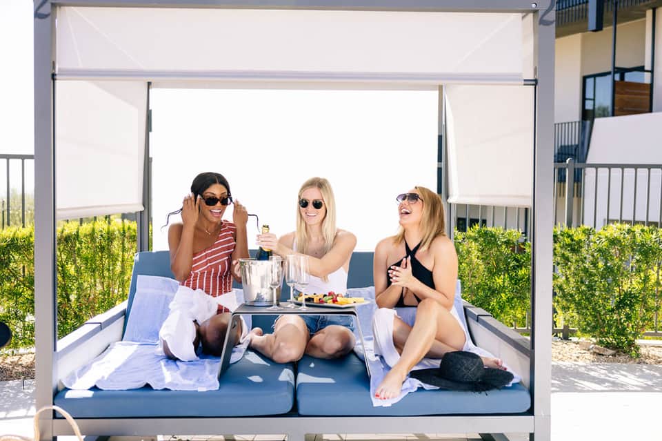 Three women relaxing on lounge bed under white pergola shade with drinks and snacks