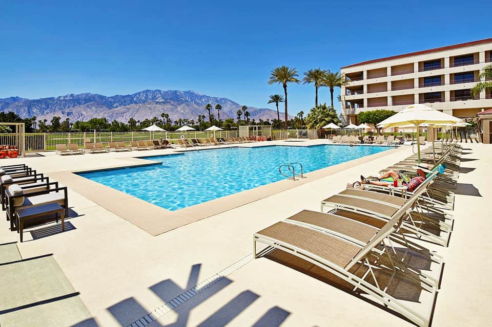 Resort pool with mountain backdrop, palm trees, lounge chairs, and multi-story hotel building