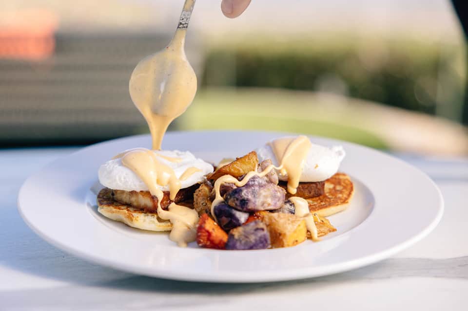 Gourmet eggs Benedict with sauce being drizzled over on white plate outdoors
