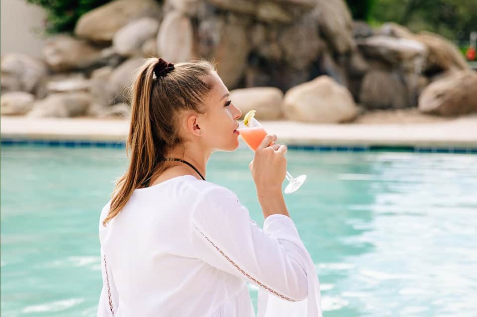 Woman in white robe enjoying cocktail poolside with stone features and tropical landscaping