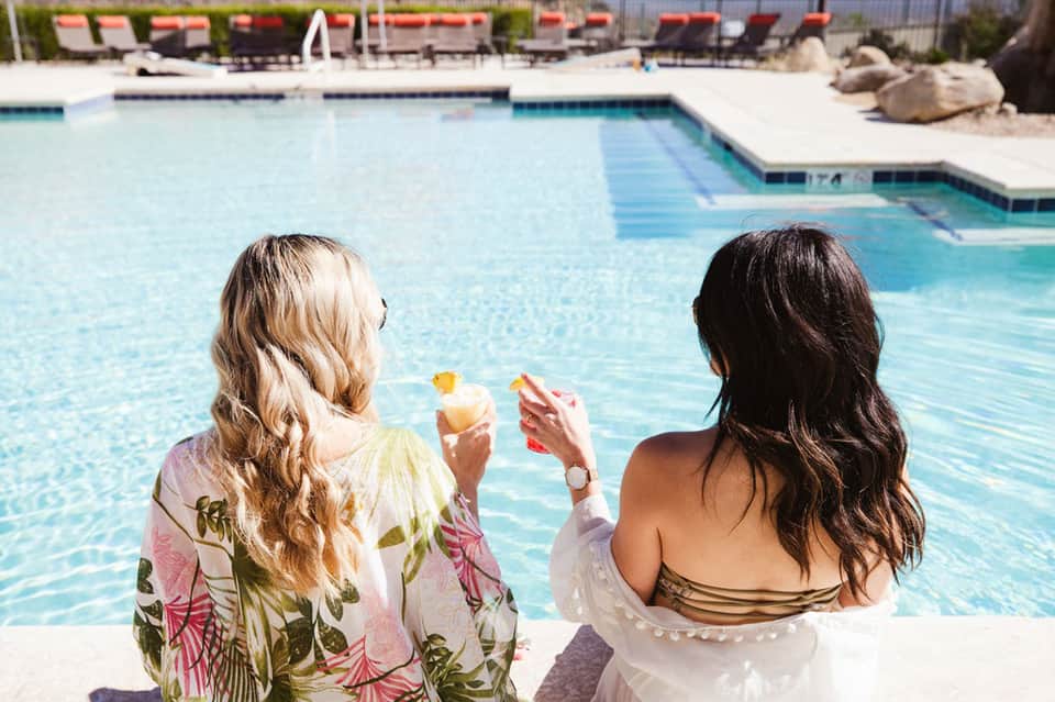 Two women at poolside holding drinks, facing crystal-blue resort swimming pool