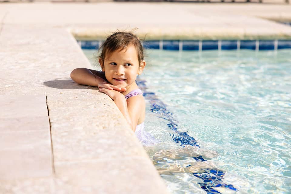 Young child in swimming pool leaning on edge, smiling at camera with resort in background