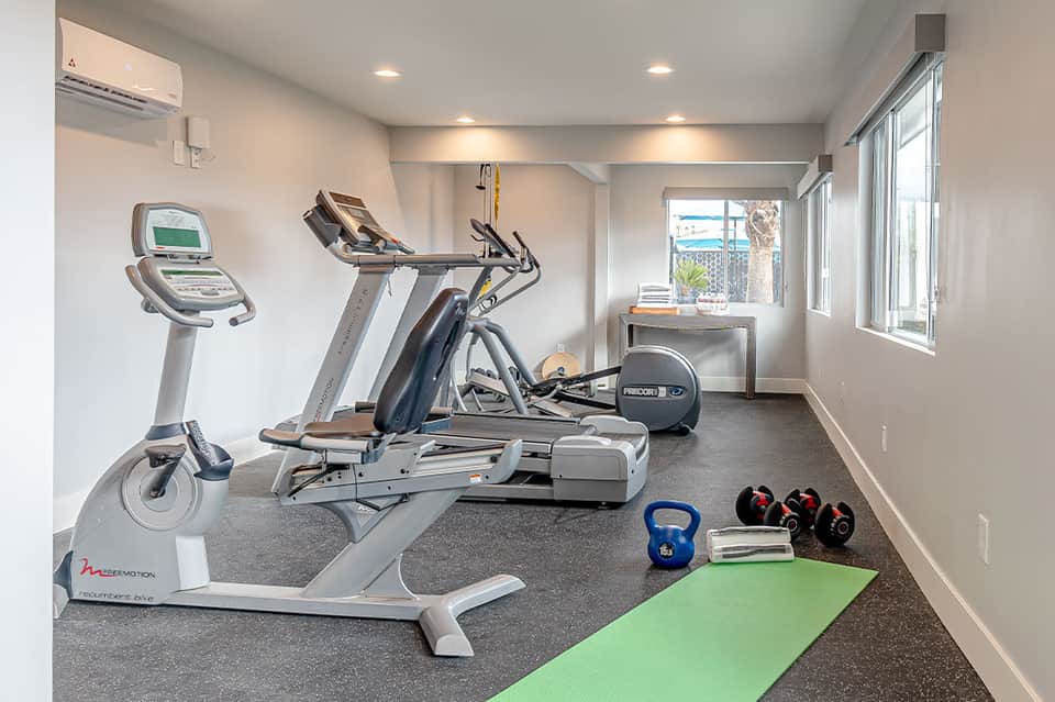 Bright fitness center with treadmill, stationary bike, elliptical machine, weights, yoga mat, and ocean view through windows