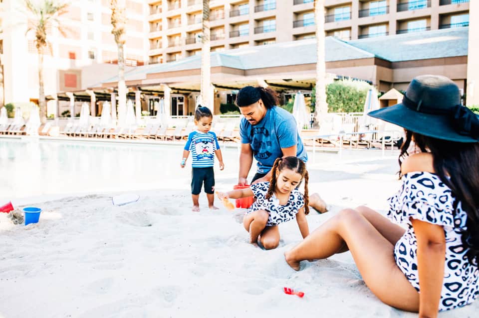 Family playing in sand on beach in front of resort with palm trees and hotel buildings