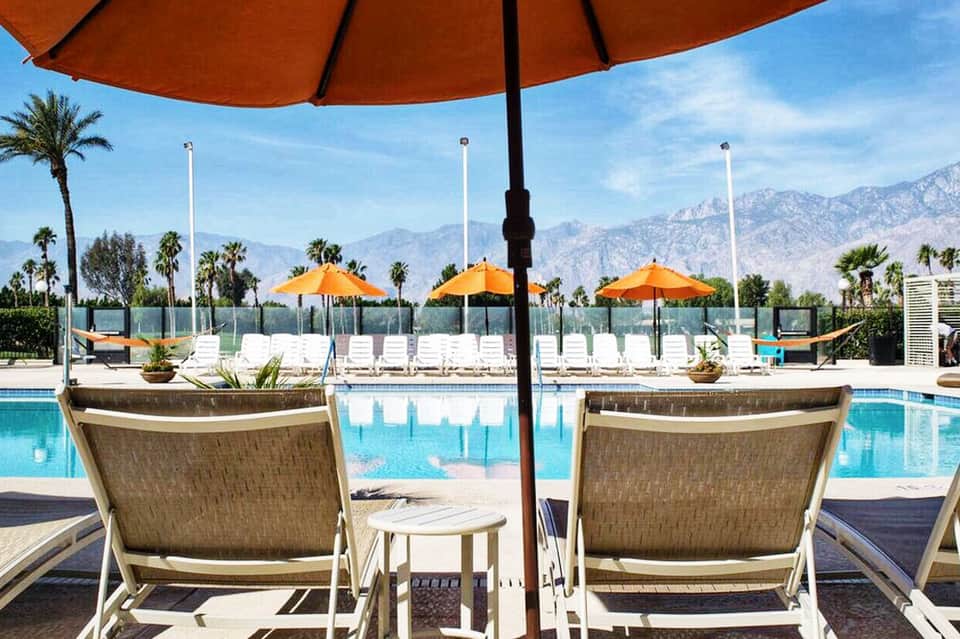 Luxury resort pool with orange umbrellas, mountain views, palm trees, and lounging chairs under shade structures
