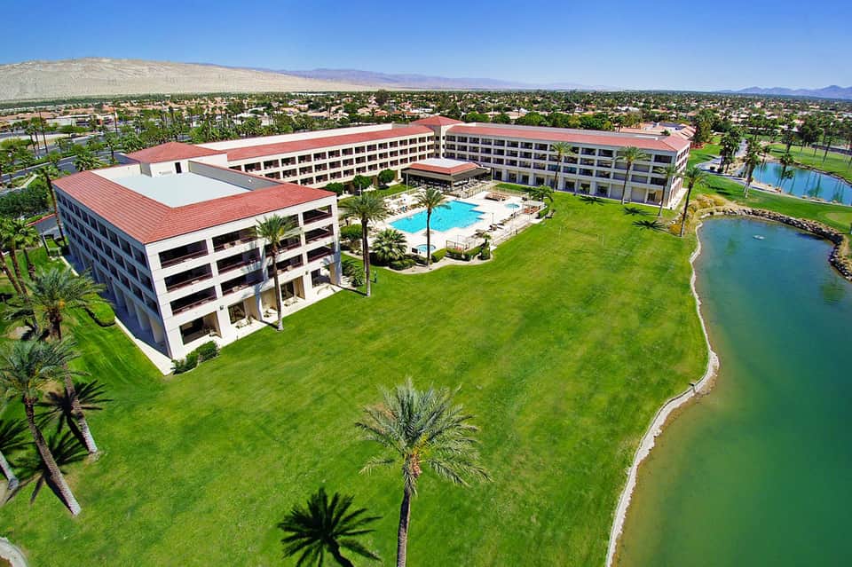 Aerial view of resort with red-roofed buildings, manicured lawns, swimming pool, and turquoise lagoon surrounded by palm trees