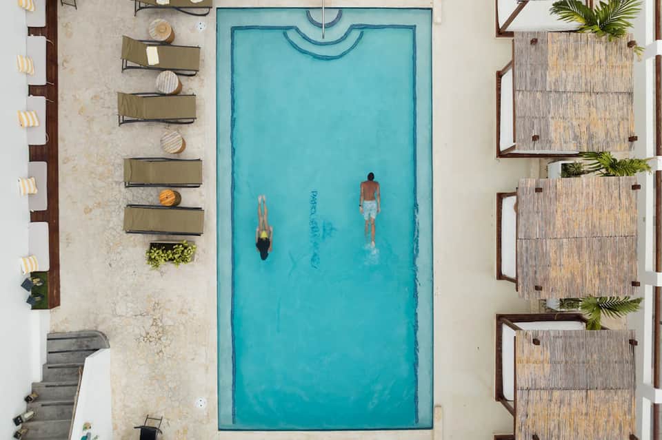 Aerial view of turquoise pool with swimmers, lounge chairs, and wooden cabanas on sides