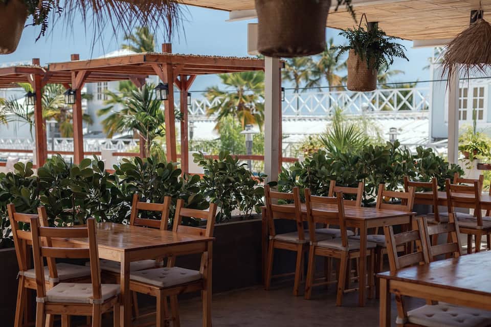 Resort terrace seating with wooden tables, chairs, hanging plants, and coastal views under pergola shade