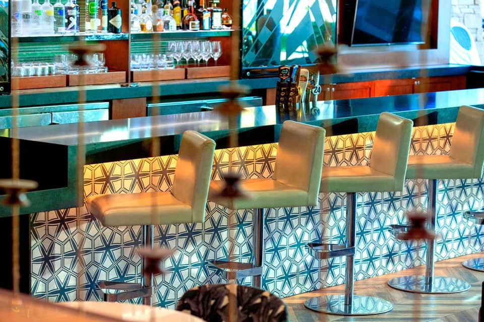 Coastal bar with turquoise and wood accents, geometric patterned bar stools, glass shelving, and ocean decor