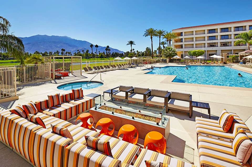 Resort pool area with striped lounge chairs, mountain backdrop, and palm trees