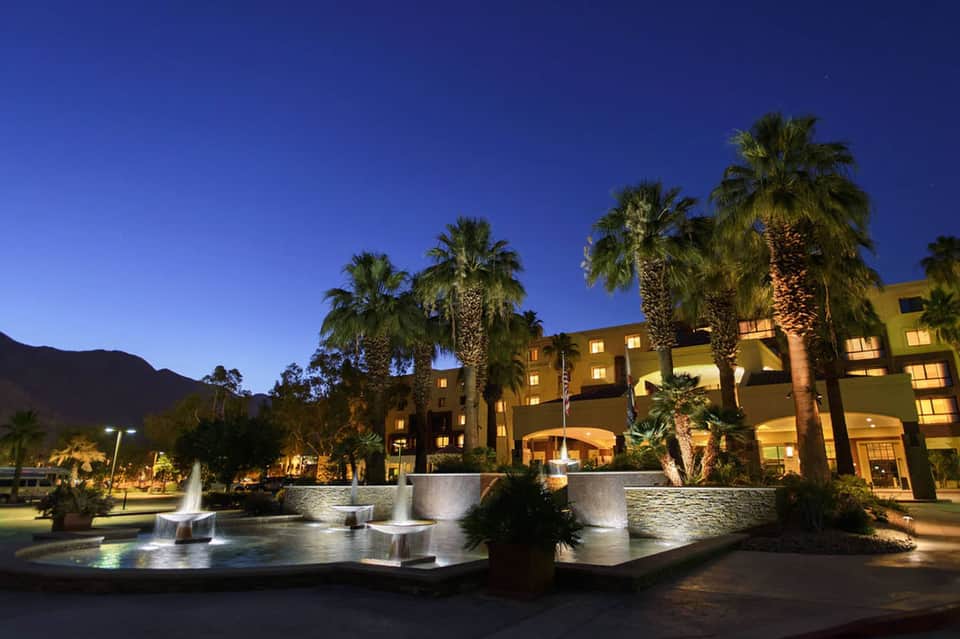 Illuminated resort courtyard at dusk with fountain, palm trees, and multi-story hotel building