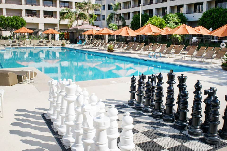 Resort pool area with orange umbrellas, lounge chairs, and giant chess pieces on deck