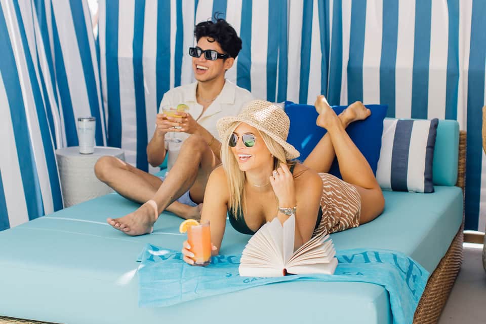 Couple relaxing on turquoise lounge by striped cabana backdrop, holding tropical drinks and sunglasses
