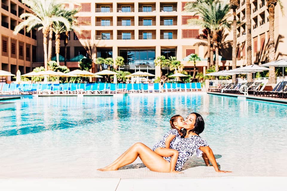 Woman and child in patterned swimsuit by resort pool with palm trees and multi-story hotel building