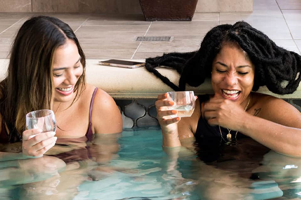 Two women relaxing in indoor thermal pool holding drinks and smiling