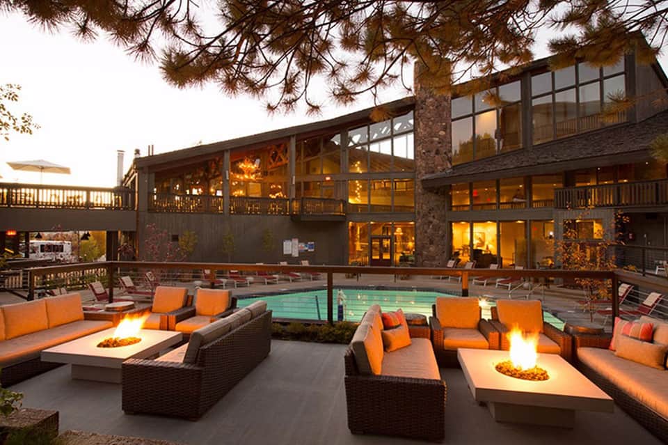 Resort pool at dusk with fire pits, orange lounge seating, multi-story building with lit windows, and tall pine tree