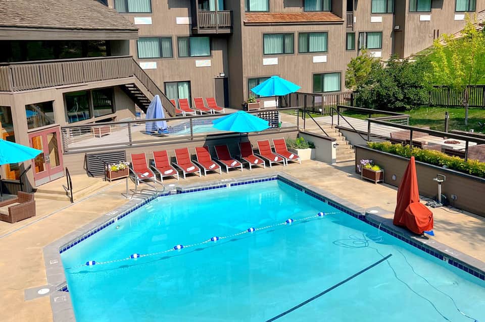 Outdoor lap pool with lane dividers, red lounge chairs, blue umbrellas, and modern building architecture