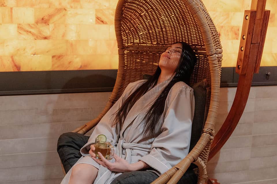 Woman relaxing in large woven egg chair holding a beverage with warm backlit wall and minimalist spa decor
