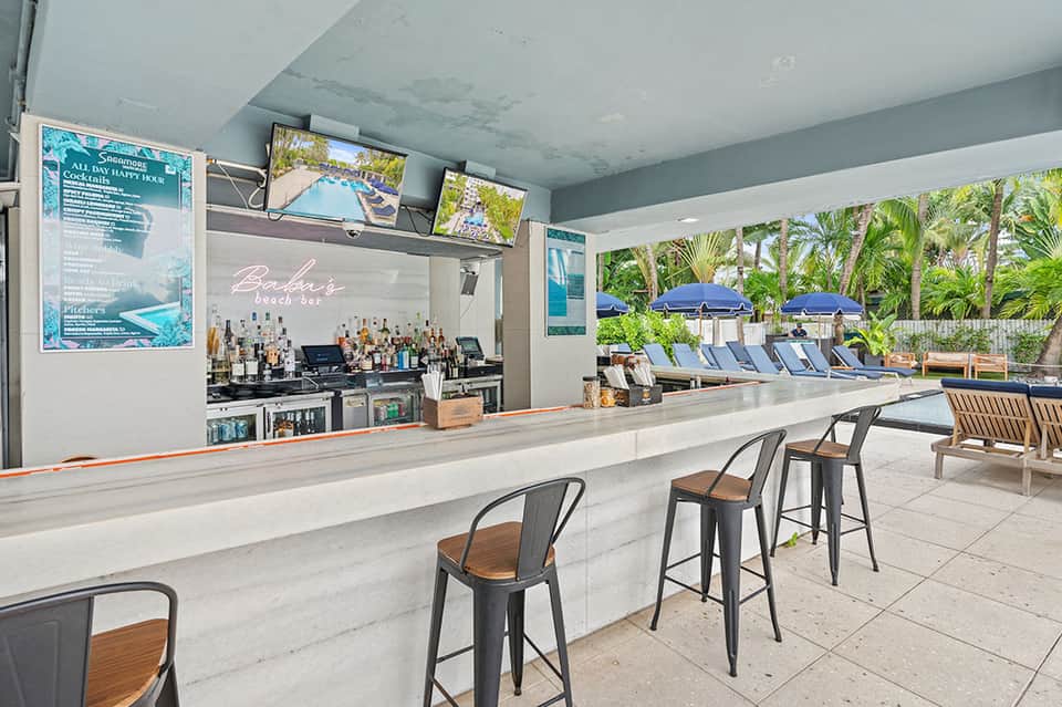 Beach bar counter with bottles and view of pool, umbrellas, and palm trees