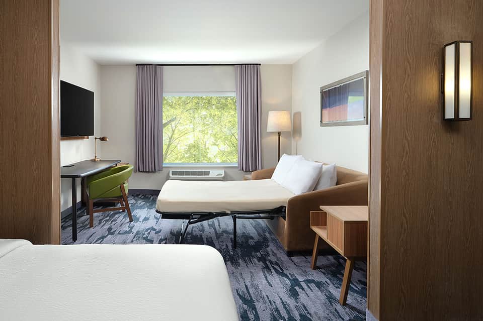 Modern hotel room with two beds, work desk, purple curtains, window view, and contemporary wood furnishings