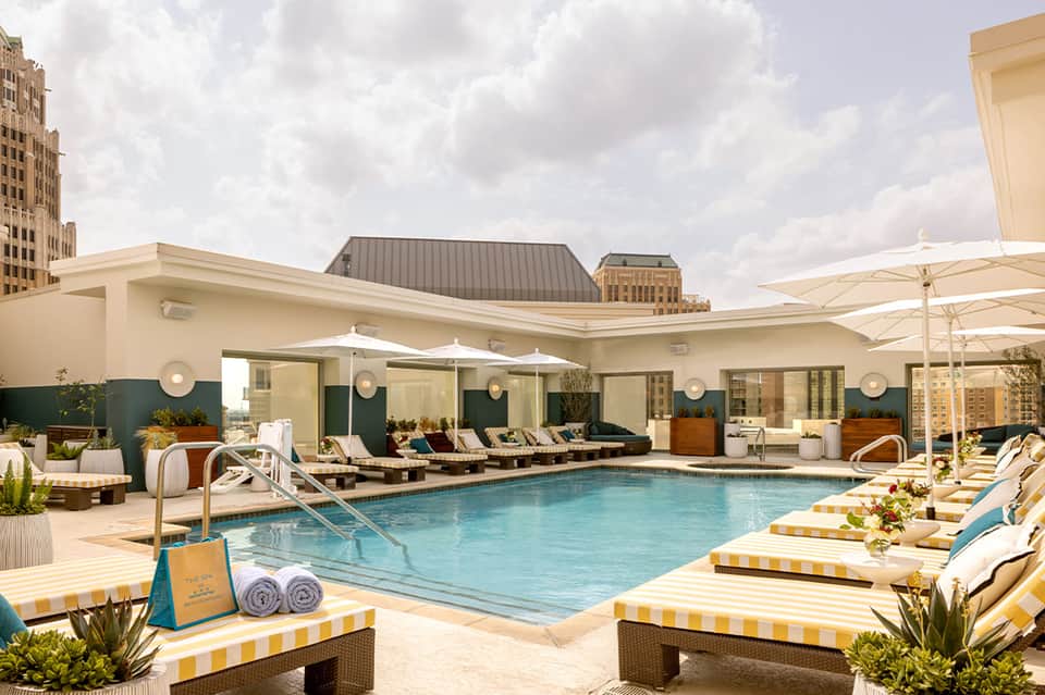 Urban rooftop pool with blue water, striped loungers, white umbrellas, and city skyline backdrop