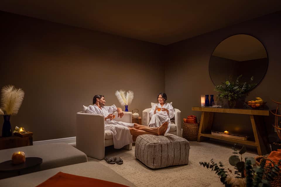 Minimalist spa wellness room with two cream seating areas, round mirror, wooden accents, ambient lighting, and neutral tones