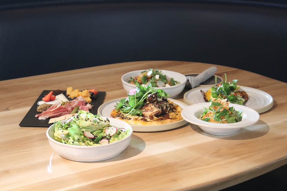 Gourmet plated dishes on wooden table including salads, cured meats, pizza, and appetizers with dark backdrop