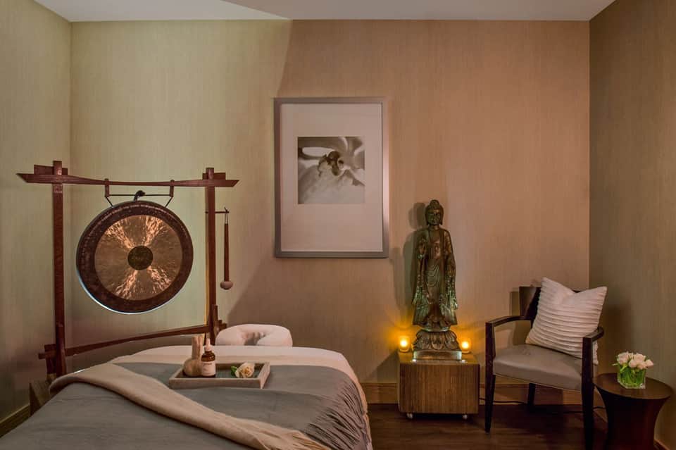 Spa treatment room with massage table, gong, Buddha statue, and warm lighting