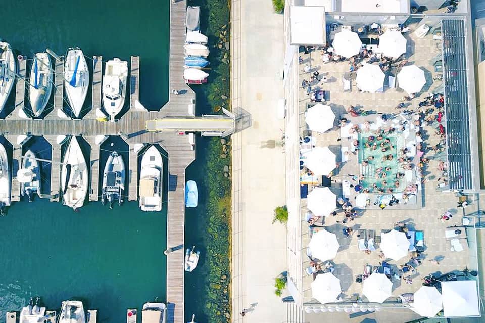 Aerial view of marina with moored boats on left, beach club with white umbrellas and pool on right
