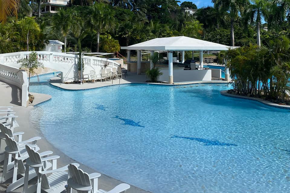 Luxury resort pool with blue water, white umbrellas, cabana, palm trees, and tropical landscaping