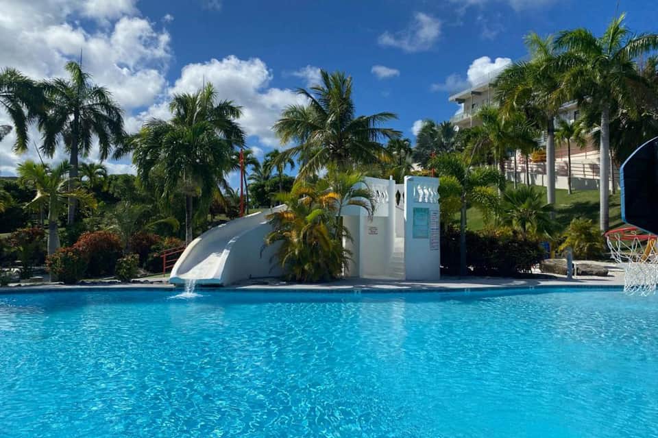 Waterslide feature in bright blue pool with palm trees and resort buildings on hillside