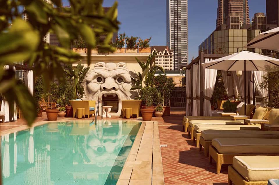 Rooftop pool with turquoise water, loungers, white umbrellas, and decorative stone lion sculpture overlooking cityscape