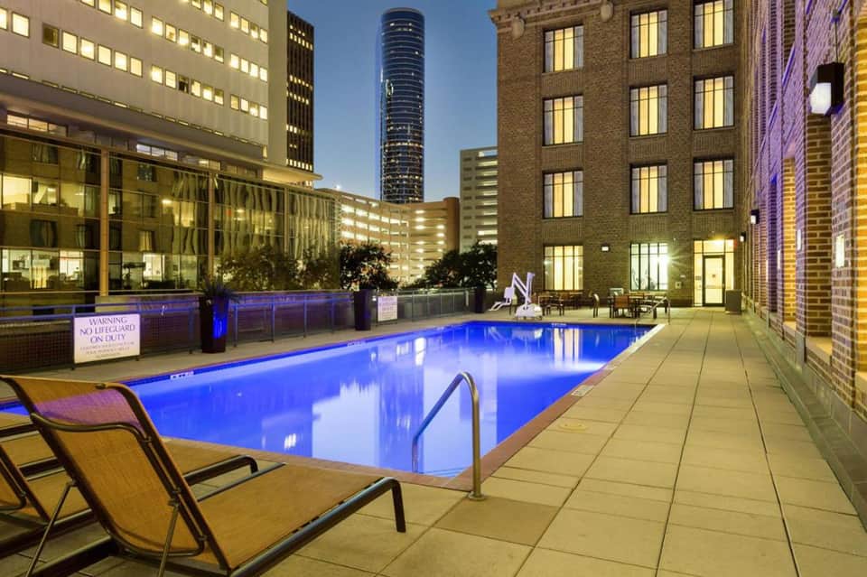 Urban rooftop swimming pool at night with blue illumination, lounge chairs, and downtown city buildings surrounding