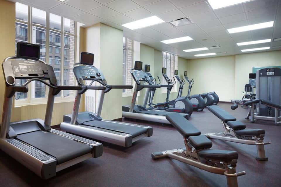 Hotel fitness center with multiple treadmills, elliptical machines, and city windows