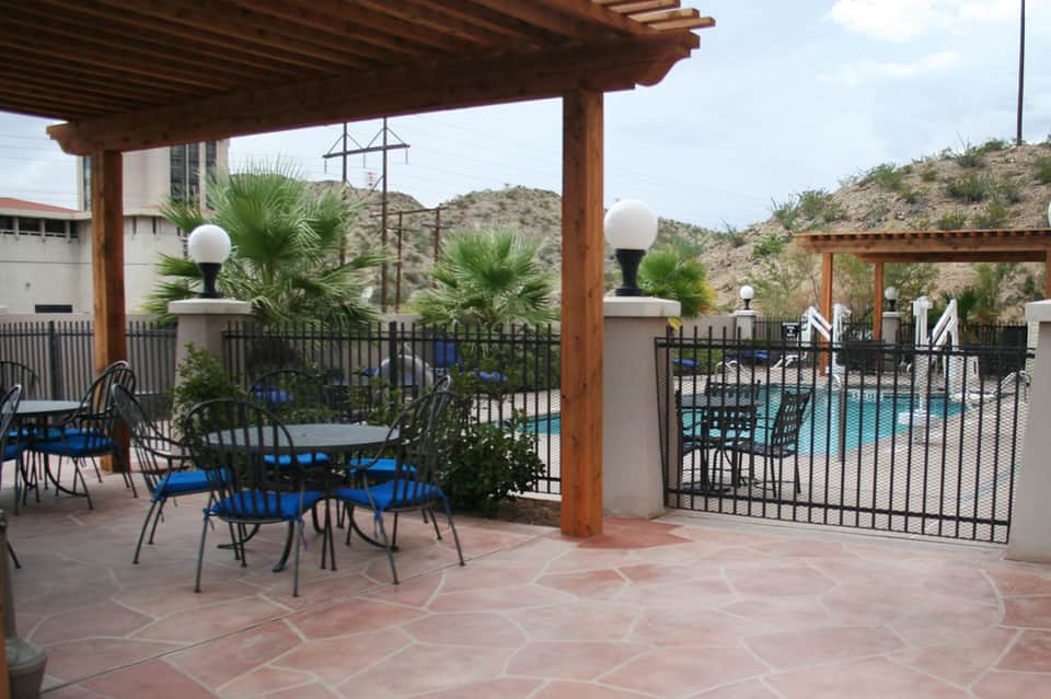 Covered patio seating area with wooden pergola, metal chairs, and pool view beyond fence