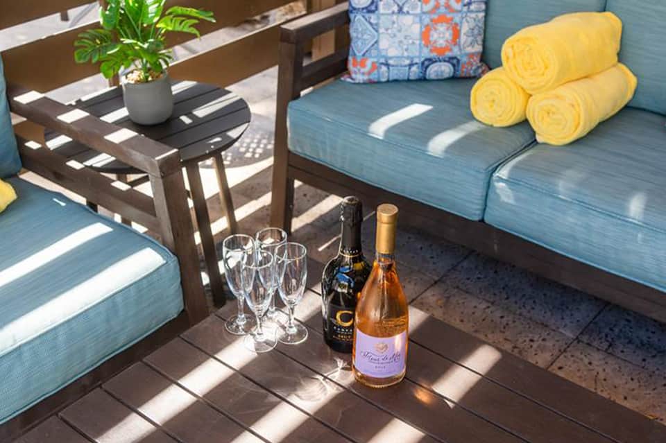 Sunny outdoor patio with blue lounge cushions, wine bottles, glasses, and rolled yellow towels
