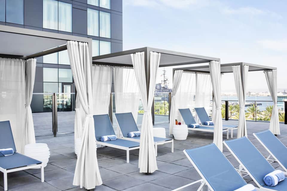 Rooftop lounge with white cabanas, blue loungers, and ocean view with palm trees and boats