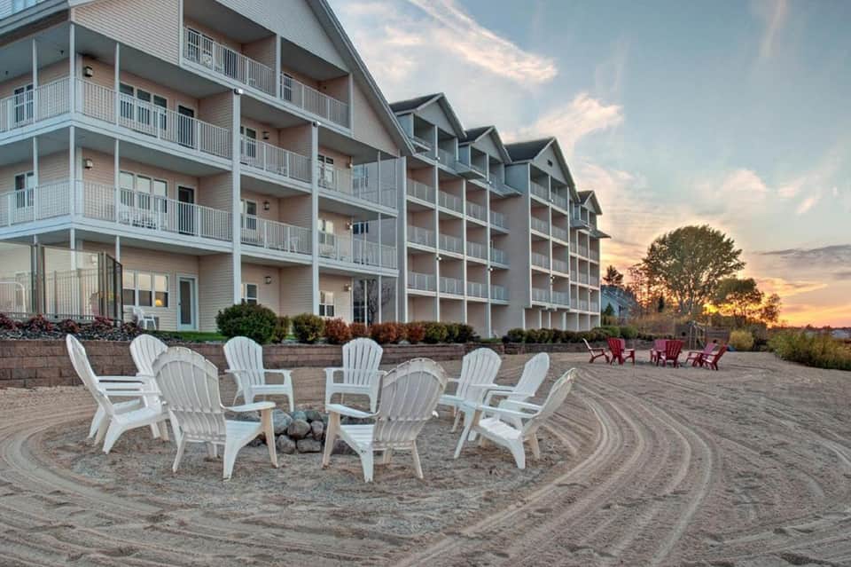 Beach bonfire seating area with white Adirondack chairs and residential buildings at sunset