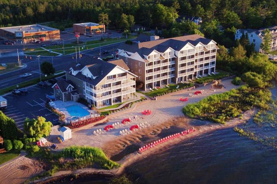 Aerial view of waterfront resort with sandy beach, red umbrellas, and forested backdrop